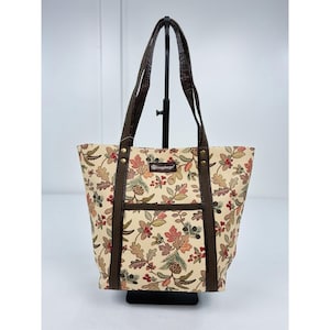 May include: A tote bag featuring a cream-colored fabric with a repeating pattern of oak leaves, pine cones, and red berries. The bag includes dark brown straps, a front pocket, and a small rectangular patch with the text Longaberger.