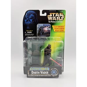 May include: A Star Wars The Power of the Force Darth Vader action figure in original packaging. The black-clad figure holds a red electronic lightsaber. The card features the Star Wars logo, a warning label, and text stating Electronic Lightsaber Really Glows.