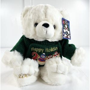 May include: A white plush teddy bear wearing a green sweater with the words "Happy Holiday" embroidered on the front. The sweater features a snowman and other holiday designs. The bear's paws have the year "2006" embroidered on them.
