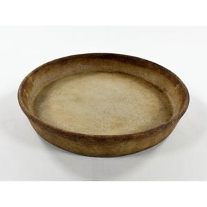 May include: A round, shallow, brown ceramic dish with a light tan interior. The dish has a slightly raised rim and a rustic, aged appearance. The dish is likely used for baking or serving.