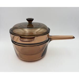 May include: A translucent amber-colored glass saucepan with a matching lid. The pot features a long, flat handle on one side and a smaller integrated handle on the opposite side. The lid has a central knob handle and fits securely over the rim.