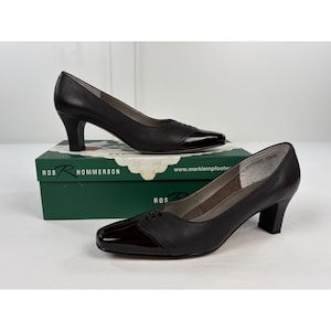 May include: A pair of dark brown leather pumps with a glossy patent leather toe cap and a mid-height heel. The shoes feature subtle stitching details on the vamp and are displayed with their original green and white Ros Hommerson brand box.