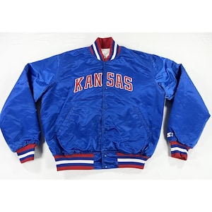 May include: A blue satin jacket with the text "KAN SAS" in white and red lettering across the chest. The jacket has a snap-button closure, red and white striped cuffs and hem, and a white and red collar.