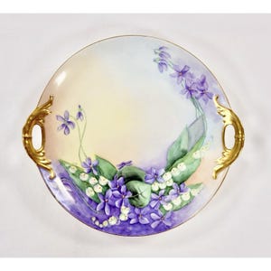 May include: A round, hand-painted porcelain serving tray with gold handles. The tray features a floral design of purple violets and white lily of the valley flowers with green leaves. The background has a gradient of yellow to purple.
