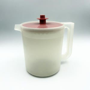 May include: A translucent white plastic pitcher featuring a cylindrical body, a sturdy side handle, and a pouring spout. The lid is a flat, circular design in a muted reddish-brown color, topped with a small, matching central knob handle.