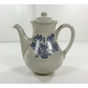 May include: A ceramic teapot with a lid and handle. The teapot is a light gray color with a blue floral design on the front. The teapot has a spout for pouring and a rounded body.