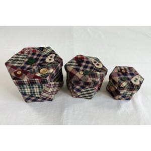 May include: Three hexagonal fabric boxes in varying sizes. The boxes are patchwork with a plaid pattern in shades of burgundy, green, and navy. Each box is adorned with decorative buttons and felt appliqués, including the number "8".