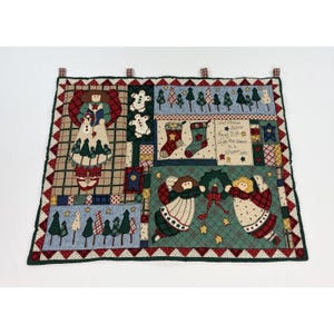 May include: A rectangular fabric wall hanging featuring a patchwork design with appliqued figures of angels, gingerbread men, Christmas stockings, and trees. The text reads: Christmas Angels Flying High Light the Stars in a Winter Sky.