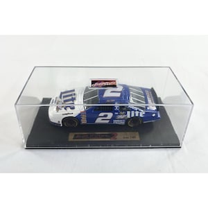 May include: A blue and white diecast race car model, number 2, displayed in a clear acrylic case. The car features sponsor logos and the name "Rusty Wallace" on a small plaque. The model is a scale of 1:43.