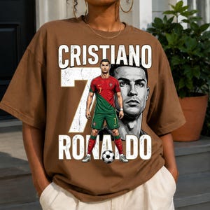 May include: A brown short-sleeve t-shirt featuring a graphic print of Cristiano Ronaldo in a red and green soccer uniform. The design includes a large white number 7, the text CRISTIANO RONALDO, and a soccer ball at the bottom of the graphic.