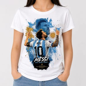 May include: A white t-shirt featuring a graphic print of Lionel Messi. The design includes a back view of the player in a blue and white striped jersey with the number 10, a portrait, a sun emblem, blue paint splatters, and the text MESSI DIOS PATRIA FAMILIA.
