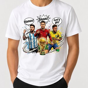 May include: A white t-shirt featuring a graphic illustration of three soccer players in their respective national team jerseys. The design includes speech bubbles with the text Vamos!, Siuuu!, and Let's go!, centered above a gold trophy.