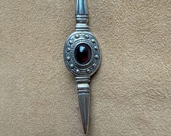 May include: A silver-toned brooch with a central oval design featuring a deep red gemstone. The brooch has a symmetrical design with pointed ends and a circular center, showcasing a vintage aesthetic.