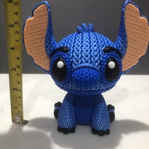 Op de afbeelding: Een blauw beeldje van een stripfiguur met grote oren en zwarte ogen. Het beeldje is ongeveer 10 cm hoog. De oren zijn perzikkleurig en het lichaam heeft een getextureerd, gebreid uiterlijk. Het personage zit met de benen naar voren.