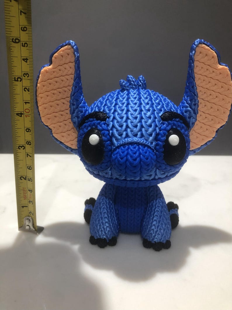 Op de afbeelding: Een blauw beeldje van een stripfiguur met grote oren en zwarte ogen. Het beeldje is ongeveer 10 cm hoog. De oren zijn perzikkleurig en het lichaam heeft een getextureerd, gebreid uiterlijk. Het personage zit met de benen naar voren.