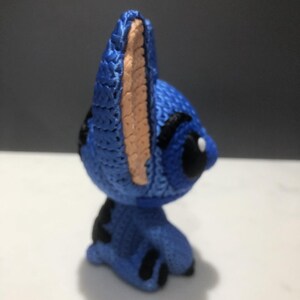 Op de afbeelding: Een blauw gehaakt figuur van een stripfiguur met grote oren en zwarte accenten. Het figuur heeft een lichtbruin binnenoor en een zwart oog. Het figuur staat op een wit oppervlak.