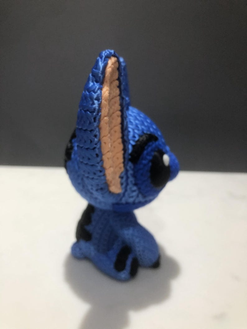 Op de afbeelding: Een blauw gehaakt figuur van een stripfiguur met grote oren en zwarte accenten. Het figuur heeft een lichtbruin binnenoor en een zwart oog. Het figuur staat op een wit oppervlak.