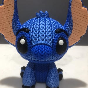 Op de afbeelding: Blauw beeldje van een stripfiguur met grote oren en zwarte ogen. Het beeldje heeft een getextureerd, gebreid uiterlijk, met lichtbruine oren en zwarte accenten op de voeten en wenkbrauwen. Het personage zit.