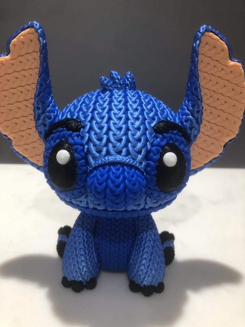 Op de afbeelding: Blauw beeldje van een stripfiguur met grote oren en zwarte ogen. Het beeldje heeft een getextureerd, gebreid uiterlijk, met lichtbruine oren en zwarte accenten op de voeten en wenkbrauwen. Het personage zit.