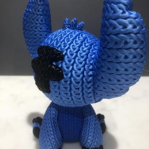 Op de afbeelding: Een blauw beeldje van een stripfiguur met grote oren en een zwarte vlek op zijn hoofd. Het beeldje heeft een getextureerd, geweven uiterlijk en staat op een wit oppervlak. De voeten en handen van het personage zijn zwart.