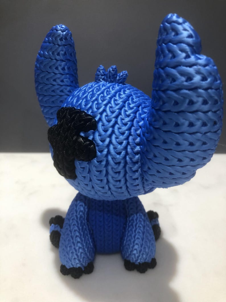 Op de afbeelding: Een blauw beeldje van een stripfiguur met grote oren en een zwarte vlek op zijn hoofd. Het beeldje heeft een getextureerd, geweven uiterlijk en staat op een wit oppervlak. De voeten en handen van het personage zijn zwart.
