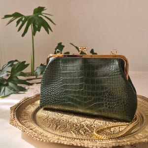 May include: A dark green, crocodile-textured clutch with a gold-toned frame and clasp. The bag features a delicate gold chain strap and rests on a decorative gold-colored tray. The clutch is a stylish accessory.