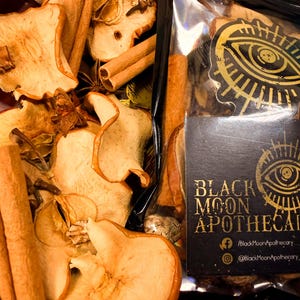 May include: A close-up shot of dried apple slices, cinnamon sticks, and star anise. A black package with gold text and an eye design reads "Black Moon Apothecary". The items are arranged in a dark red bowl.