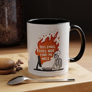 May include: White ceramic mug with a black interior and handle. The mug features a graphic of a skeleton at a computer with flames and the text "THIS EMAIL DOES NOT FIND ME WELL". A spoon and a pastry are in the foreground.