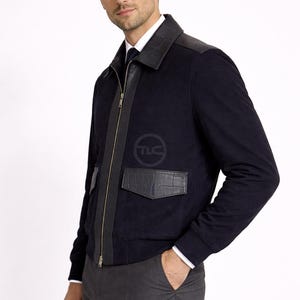 May include: A navy blue jacket with leather detailing on the shoulders, pockets, and zip. The jacket has a full-length zip and a logo on the chest. Worn with a white shirt and grey trousers.