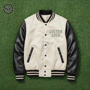 May include: Cream-colored varsity jacket with black sleeves. The jacket features a black and white striped collar, cuffs, and hem. The text "CUSTOM LOGO" is printed on the front. The jacket has two front pockets and a button-up closure. The jacket is displayed on a green surface.