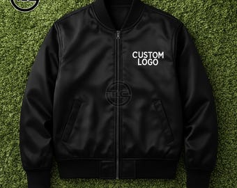 May include: Black bomber jacket with a zipper closure, two front pockets, and a ribbed collar and cuffs. The jacket features the text "CUSTOM LOGO" in white on the chest. The jacket is displayed on a green surface.