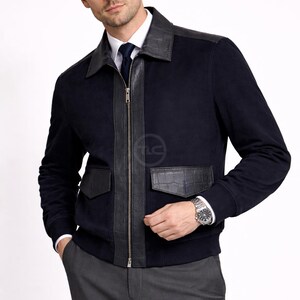 May include: Navy blue jacket with black leather detailing. It has a zip closure, leather shoulder panels, and two front pockets. The jacket is worn over a white shirt and grey trousers. A silver watch is visible.