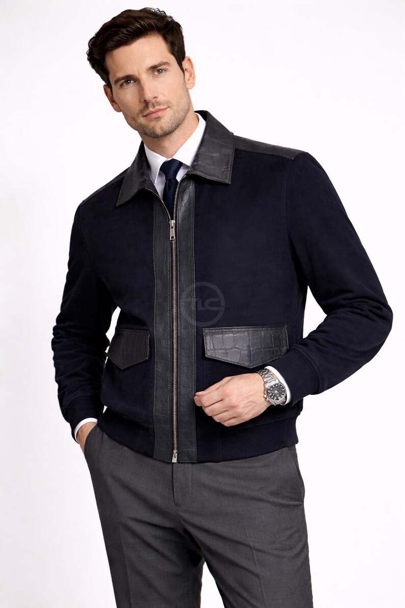 May include: Navy blue jacket with black leather detailing. It has a zip closure, leather shoulder panels, and two front pockets. The jacket is worn over a white shirt and grey trousers. A silver watch is visible.