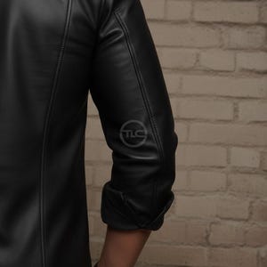 Puede incluir: Chaqueta de cuero negra con una textura sutil. La chaqueta presenta un dise&ntilde;o cl&aacute;sico con una parte delantera abotonada y un peque&ntilde;o logotipo en la manga. Las mangas est&aacute;n enrolladas, revelando el antebrazo. La chaqueta est&aacute; sobre una pared de ladrillos.