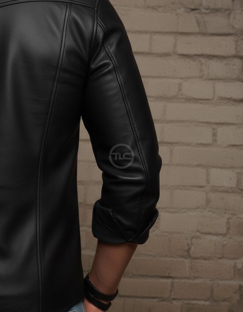 Puede incluir: Chaqueta de cuero negra con una textura sutil. La chaqueta presenta un dise&ntilde;o cl&aacute;sico con una parte delantera abotonada y un peque&ntilde;o logotipo en la manga. Las mangas est&aacute;n enrolladas, revelando el antebrazo. La chaqueta est&aacute; sobre una pared de ladrillos.