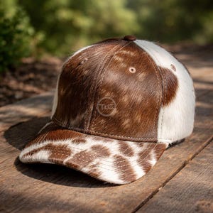 May include: A baseball cap with a brown and white cowhide pattern. The cap has a curved peak and a brown leather button on top. The logo "TGC" is visible on the front. The hat is made of a soft, textured material.