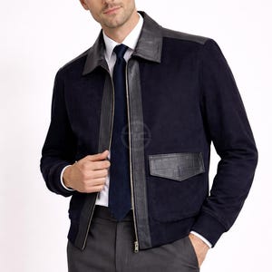 May include: A navy blue suede jacket with black leather detailing on the collar, shoulders, and pockets. The jacket has a front zip and is worn with a white shirt, a navy tie, and grey trousers.