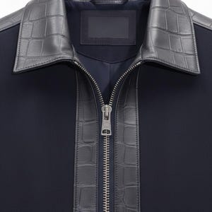 May include: A navy blue jacket with a dark grey, textured faux-leather collar and trim. The jacket has a full-length silver zip and a small, rectangular label. The design is modern and stylish, suitable for various occasions.