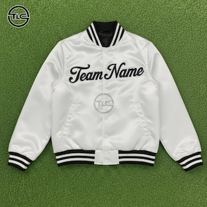 May include: White satin varsity jacket with black and white striped trim on the collar, cuffs, and hem. The jacket features the text "Team Name" in black script across the chest. A logo is visible on the upper left.
