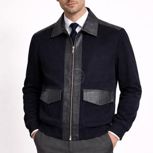 May include: A navy blue jacket with black leather detailing on the shoulders, front, and pockets. It features a zip closure and two front pockets. The model is wearing a white shirt and grey trousers.