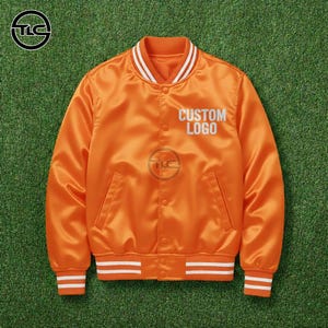 May include: Bright orange satin bomber jacket with white striped detailing on the collar, cuffs, and hem. The jacket has a button-up front, two pockets, and the text "CUSTOM LOGO" in white. It's shown on a green surface.