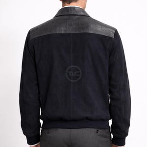 May include: A navy blue suede jacket with a black leather shoulder panel. The jacket has a ribbed hem and cuffs, and a classic collar. The model is wearing grey trousers and a white shirt.