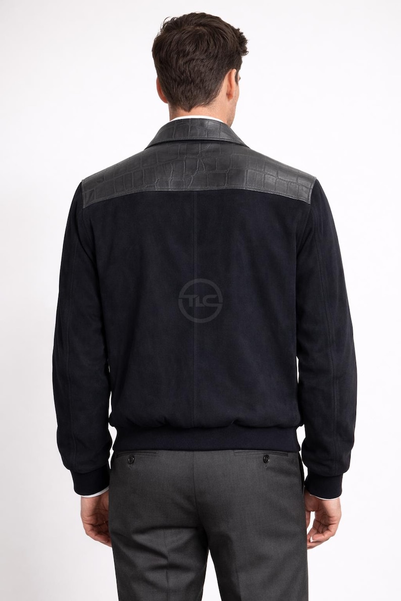 May include: A navy blue suede jacket with a black leather shoulder panel. The jacket has a ribbed hem and cuffs, and a classic collar. The model is wearing grey trousers and a white shirt.