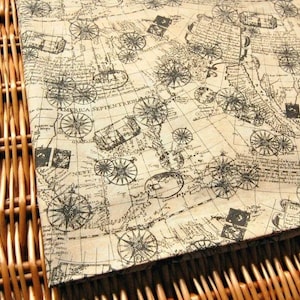 May include: A tan fabric printed with a vintage map design. The map includes compass roses, cartouches, and the text "AMERICA SEPTENTRIONA". The fabric is folded, showing the woven texture. The overall style evokes antique maps.