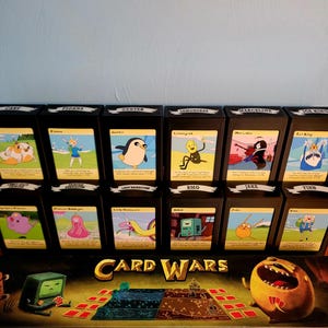 Può includere: Un set di gioco da tavolo Adventure Time Card Wars Ultimate Collection con dodici mazzi di carte a tema personaggio disposti in due file sopra la scatola principale, che mostra il titolo Card Wars e l'illustrazione del tabellone.
