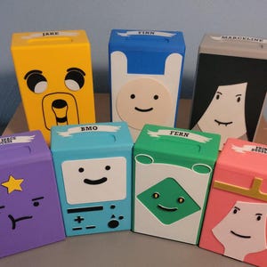 Può includere: Sette scatole rettangolari colorate con personaggi di Adventure Time. Ogni scatola ha un design diverso per i personaggi, tra cui Jake, Finn, Marceline, Lumpy Space Princess, BMO, Fern e Princess Bubblegum. Le scatole sono gialle, blu, nere, viola, verde acqua, verdi e rosa.