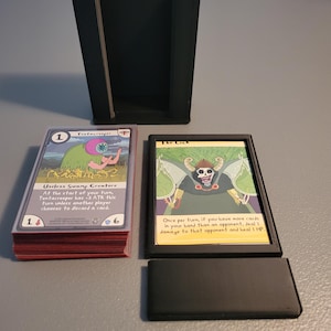 Può includere: Portacarte nero con due carte collezionabili e un mazzo di carte. Le carte presentano illustrazioni e testo. Una carta è etichettata "Tentacreeper" e l'altra "The Lich". Il mazzo di carte è rosso e il portacarte è nero.