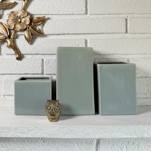 May include: A gold-colored floral wall hanging and three square, light-green ceramic vases are displayed on a white shelf. A small, gold-colored owl figurine sits in front of the vases. The background is a white brick wall.