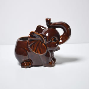 May include: A glossy dark brown ceramic elephant figurine with a hollow center, featuring large ears, a raised trunk, and a smooth, reflective finish. The elephant is depicted in a seated position with visible textured details on the ears and feet.
