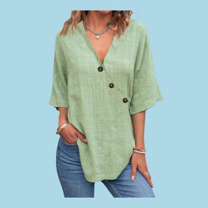 May include: A light green, V-neck blouse with three brown buttons on the side. The blouse has short sleeves and a textured fabric. The model is wearing blue jeans and accessories.